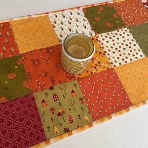 May include: A rectangular patchwork table runner featuring a variety of fabric squares in shades of orange, olive green, red, and cream. The patterns include pumpkins, houses, corn, and geometric shapes. A glass candle holder sits on the runner.