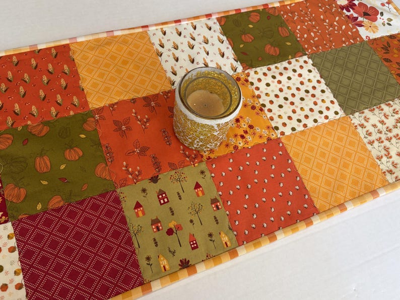 May include: A rectangular patchwork table runner featuring a variety of fabric squares in shades of orange, olive green, red, and cream. The patterns include pumpkins, houses, corn, and geometric shapes. A glass candle holder sits on the runner.