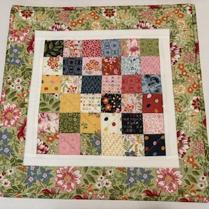 May include: A square quilted fabric piece featuring a central grid of thirty-six small, multi-colored patterned squares framed by a white border and an outer border of green fabric decorated with a dense floral print of red, pink, and blue flowers.