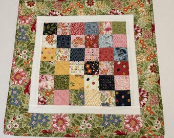 May include: A square quilted fabric piece featuring a central grid of thirty-six small, multi-colored patterned squares framed by a white border and an outer border of green fabric decorated with a dense floral print of red, pink, and blue flowers.