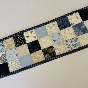 May include: A rectangular quilted table runner featuring a patchwork design of squares in shades of navy blue, light blue, cream, and white. The fabric squares display patterns of birds, trees, leaves, and cursive script text with a dark blue border.