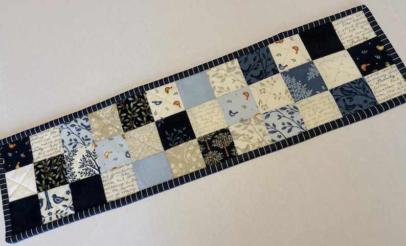 May include: A rectangular quilted table runner featuring a patchwork design of squares in shades of navy blue, light blue, cream, and white. The fabric squares display patterns of birds, trees, leaves, and cursive script text with a dark blue border.