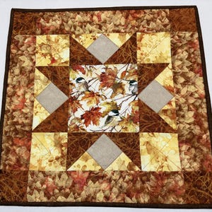 May include: A square fabric quilt featuring a central panel with chickadees perched among autumn leaves. The design includes a star pattern with brown, tan, and yellow leaf-print fabric, accented by solid light gray diamond-shaped patches.