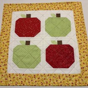 May include: A quilted placemat featuring four apple designs. Two red apples with white polka dots and swirl patterns, and two green apples with white polka dots. The placemat has a yellow floral border and white background.