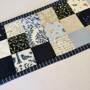 May include: A rectangular quilted table runner featuring a patchwork design of navy blue, light blue, and cream fabric squares. The pattern includes botanical leaf motifs, small bird illustrations, and script text. It is finished with a navy blue striped border.