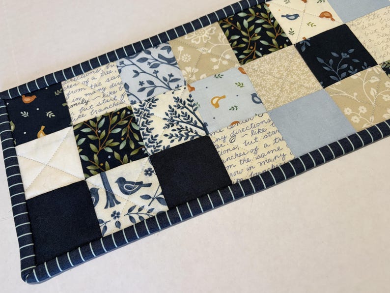 May include: A rectangular quilted table runner featuring a patchwork design of navy blue, light blue, and cream fabric squares. The pattern includes botanical leaf motifs, small bird illustrations, and script text. It is finished with a navy blue striped border.