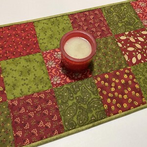 Puede incluir: Camino de mesa acolchado con dise&ntilde;o de patchwork en tonos rojos y verdes. Un portavelas rojo con una vela blanca se encuentra sobre el camino. El camino presenta varios patrones florales y de cachemira.