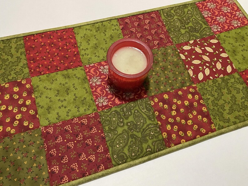 Puede incluir: Camino de mesa acolchado con dise&ntilde;o de patchwork en tonos rojos y verdes. Un portavelas rojo con una vela blanca se encuentra sobre el camino. El camino presenta varios patrones florales y de cachemira.