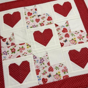 May include: A handmade Valentine's Day themed quilt featuring a repeating pattern of red polka-dot hearts and cat-shaped blocks. The cat blocks have a floral and heart pattern. The quilt has a red polka-dot border.