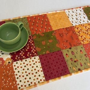 May include: A patchwork table runner featuring a variety of autumn-themed fabric squares in shades of orange, red, green, and white, with patterns including pumpkins, leaves, and houses. A solid light green ceramic teacup and saucer sit on top.