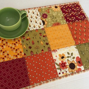 Peut inclure: Un set de table rectangulaire en tissu matelass&eacute; pr&eacute;sentant un motif patchwork aux th&egrave;mes automnaux dans des tons orange, rouge, jaune et vert, surmont&eacute; d'une tasse &agrave; th&eacute; et d'une soucoupe en c&eacute;ramique vert clair uni.