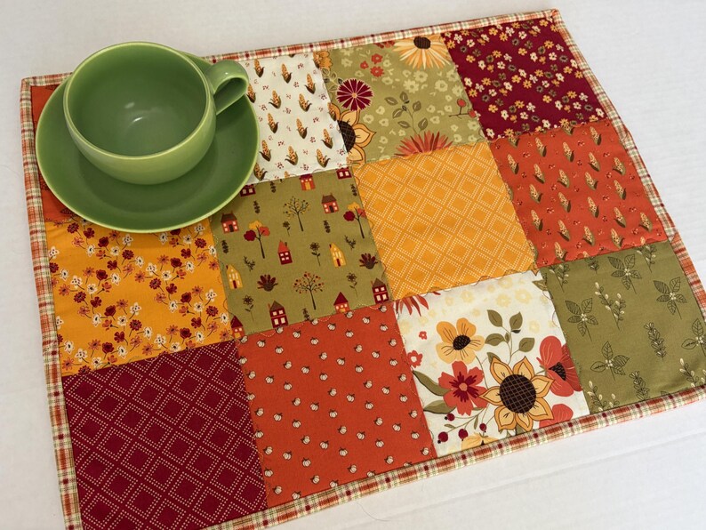 Peut inclure: Un set de table rectangulaire en tissu matelass&eacute; pr&eacute;sentant un motif patchwork aux th&egrave;mes automnaux dans des tons orange, rouge, jaune et vert, surmont&eacute; d'une tasse &agrave; th&eacute; et d'une soucoupe en c&eacute;ramique vert clair uni.