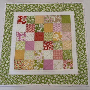 May include: A square fabric quilt featuring a central grid of multi-colored patchwork squares in shades of red, pink, yellow, and green. The center is framed by a solid white border, surrounded by an outer border of green floral fabric.