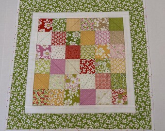 May include: A square fabric quilt featuring a central grid of multi-colored patchwork squares in shades of red, pink, yellow, and green. The center is framed by a solid white border, surrounded by an outer border of green floral fabric.