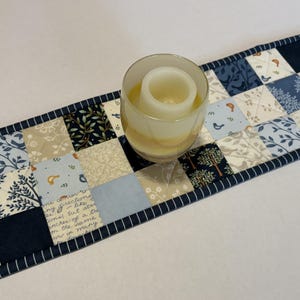 May include: A rectangular quilted table runner featuring a patchwork pattern of navy blue, light blue, and cream fabric squares with printed motifs of birds, trees, and cursive script text. A clear glass candle holder sits on the runner.