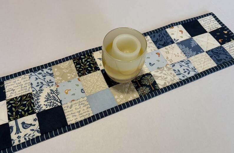 May include: A rectangular quilted table runner featuring a patchwork pattern of navy blue, light blue, and cream fabric squares with printed motifs of birds, trees, and cursive script text. A clear glass candle holder sits on the runner.