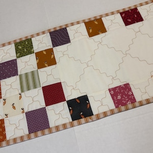 May include: A rectangular quilted table runner featuring a patchwork design of various colored fabric squares including brown, green, purple, orange, and red patterns, set against a cream background with a continuous wavy stitched grid pattern.