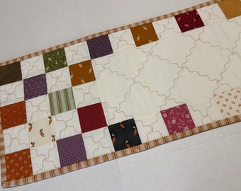 May include: A rectangular quilted table runner featuring a patchwork design of various colored fabric squares including brown, green, purple, orange, and red patterns, set against a cream background with a continuous wavy stitched grid pattern.