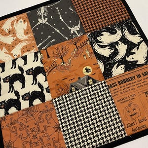May include: A Halloween-themed patchwork square with a black border. The fabric squares feature ghosts, skeletons, black cats, and a houndstooth pattern. One square has text that reads "MASS ROBBERY IN SALEM" and "MISSING".