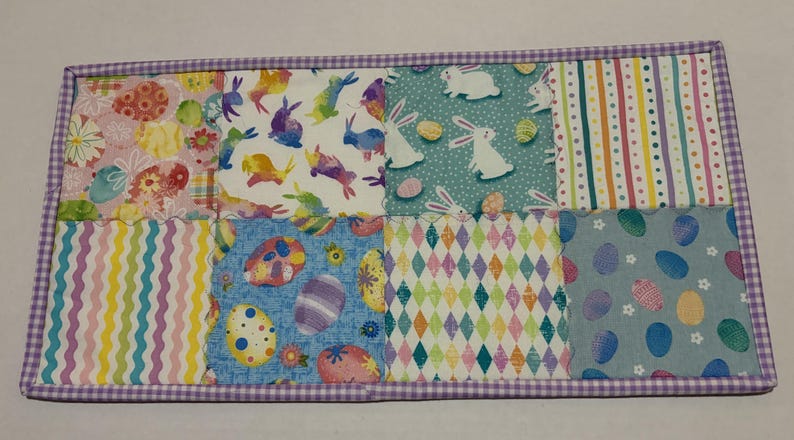 Może przedstawiać: Prostokątna podkładka pod talerz o tematyce wielkanocnej z patchworkiem. Patchwork zawiera wzory pisanek, kr&oacute;liczk&oacute;w i kolorowych kształt&oacute;w geometrycznych. Obramowanie to fioletowo-biały wz&oacute;r w kratę vichy.