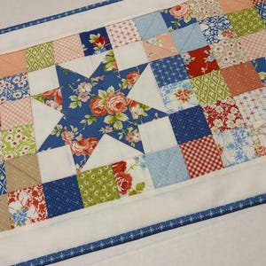 May include: A quilted table runner featuring a central blue star with floral patterns, surrounded by a patchwork of squares in various colors and patterns, including floral, gingham, and solid fabrics. The edges are finished with a white border and a blue patterned trim.