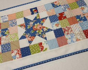 May include: A quilted table runner featuring a central blue star with floral patterns, surrounded by a patchwork of squares in various colors and patterns, including floral, gingham, and solid fabrics. The edges are finished with a white border and a blue patterned trim.