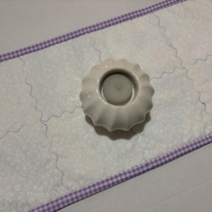 Może przedstawiać: Biały ceramiczny świecznik ze świecą stoi na białym, pikowanym bieżniku. Bieżnik ma kwiatowy wz&oacute;r i fioletową obw&oacute;dkę w kratkę vichy. Bieżnik ma około 30 cm szerokości.