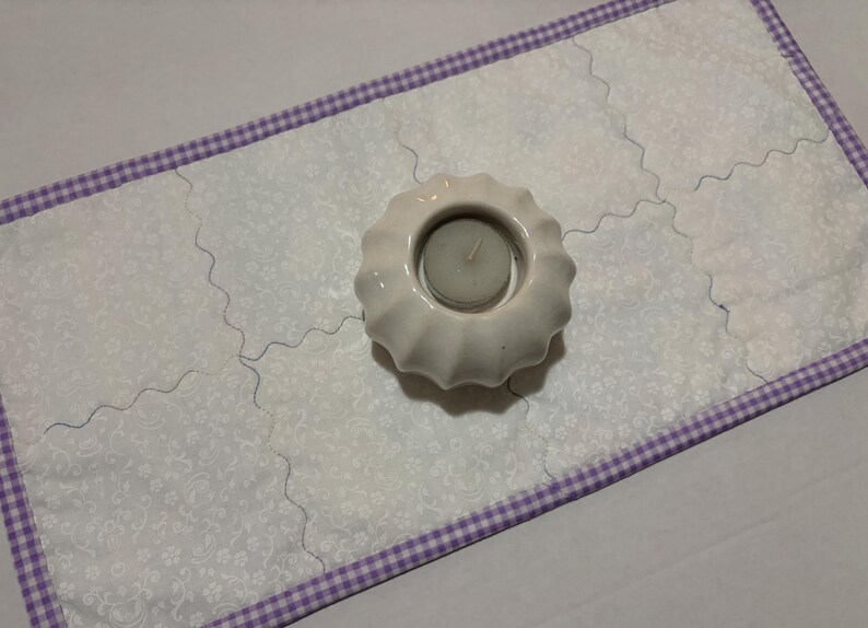 Może przedstawiać: Biały ceramiczny świecznik ze świecą stoi na białym, pikowanym bieżniku. Bieżnik ma kwiatowy wz&oacute;r i fioletową obw&oacute;dkę w kratkę vichy. Bieżnik ma około 30 cm szerokości.