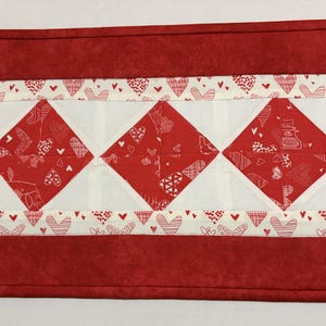 May include: A rectangular red table runner with a white and red heart pattern. The center features three red diamond shapes with white heart designs. The border is red with a white heart pattern.