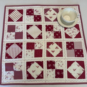 May include: A square quilted fabric mat featuring a patchwork design of alternating solid and patterned blocks in shades of deep red and white. The mat includes motifs of bees, birds, and floral patterns, with a white teacup and saucer placed on top.