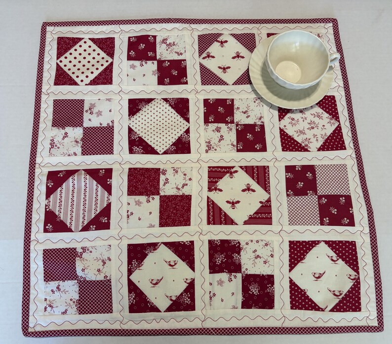 May include: A square quilted fabric mat featuring a patchwork design of alternating solid and patterned blocks in shades of deep red and white. The mat includes motifs of bees, birds, and floral patterns, with a white teacup and saucer placed on top.