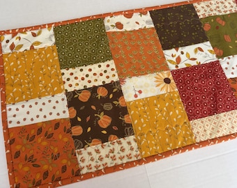 May include: A rectangular quilted table runner featuring a patchwork design of autumn-themed fabric squares. The pattern includes various prints of pumpkins, leaves, sunflowers, and berries in a color palette of orange, brown, yellow, and cream.
