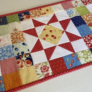 May include: A colorful patchwork table runner with a red polka dot border. The runner features squares of various floral and patterned fabrics, including cherries, flowers, and gingham. White triangles and a central starburst design add visual interest.