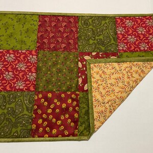 Puede incluir: Un camino de mesa de patchwork con cuadrados rojos y verdes con estampados florales y de cachemira. El camino de mesa tiene un borde verde y un reverso beige con un dise&ntilde;o de hojas. Una esquina est&aacute; doblada.