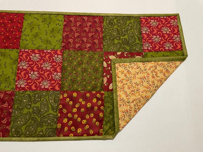 Puede incluir: Un camino de mesa de patchwork con cuadrados rojos y verdes con estampados florales y de cachemira. El camino de mesa tiene un borde verde y un reverso beige con un dise&ntilde;o de hojas. Una esquina est&aacute; doblada.