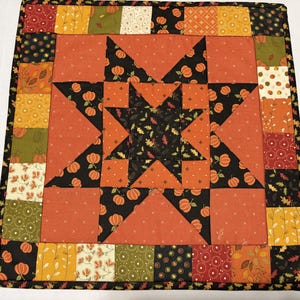 May include: A square quilted wall hanging featuring a central star pattern surrounded by a border of patchwork squares. The fabric displays autumn-themed prints including orange pumpkins, brown leaves, and floral motifs in shades of black, orange, and green.