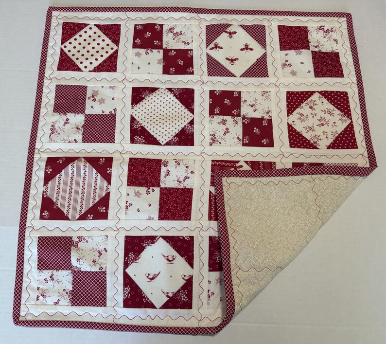 May include: A square patchwork quilt featuring a grid of alternating blocks in deep red and cream. The design includes patterns of polka dots, floral prints, bees, and birds. The quilt is finished with a red and white checkered border.