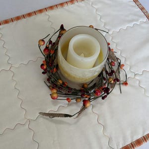 Peut inclure: Un chemin de table matelass&eacute; de couleur cr&egrave;me avec une bordure &agrave; carreaux orange. Au centre se trouve un bougeoir en verre contenant une bougie pilier blanche, entour&eacute; d'une couronne d&eacute;corative de brindilles brunes, baies rouges et bl&eacute;.