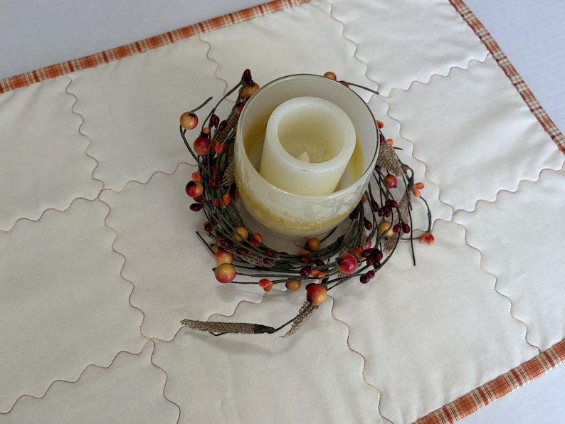 Peut inclure: Un chemin de table matelass&eacute; de couleur cr&egrave;me avec une bordure &agrave; carreaux orange. Au centre se trouve un bougeoir en verre contenant une bougie pilier blanche, entour&eacute; d'une couronne d&eacute;corative de brindilles brunes, baies rouges et bl&eacute;.