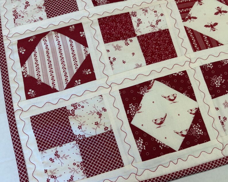 May include: A patchwork quilt featuring a repeating pattern of cream and deep red fabric squares. The design includes blocks with floral prints, stripes, and motifs of birds and bees. Each square is separated by cream sashing with red rickrack.