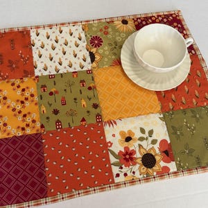 Peut inclure: Un set de table rectangulaire en patchwork compos&eacute; de carr&eacute;s de tissu aux motifs automnaux dans des tons orange, jaune, vert et bordeaux, incluant des fleurs, des feuilles et des maisons, surmont&eacute; d'une tasse et d'une soucoupe en c&eacute;ramique blanche.