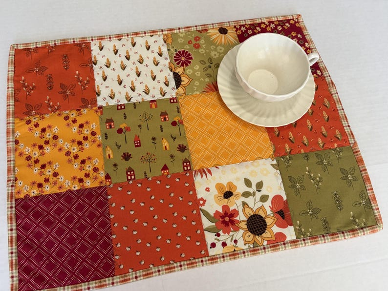 Peut inclure: Un set de table rectangulaire en patchwork compos&eacute; de carr&eacute;s de tissu aux motifs automnaux dans des tons orange, jaune, vert et bordeaux, incluant des fleurs, des feuilles et des maisons, surmont&eacute; d'une tasse et d'une soucoupe en c&eacute;ramique blanche.