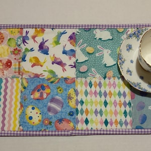 Może przedstawiać: Kolorowa podkładka na st&oacute;ł w tematyce wielkanocnej z filiżanką i spodkiem. Podkładka ma kwadraty patchworkowe z pisankami, kr&oacute;liczkami i pastelowymi wzorami. Filiżanka i spodek mają niebieski kwiatowy wz&oacute;r.