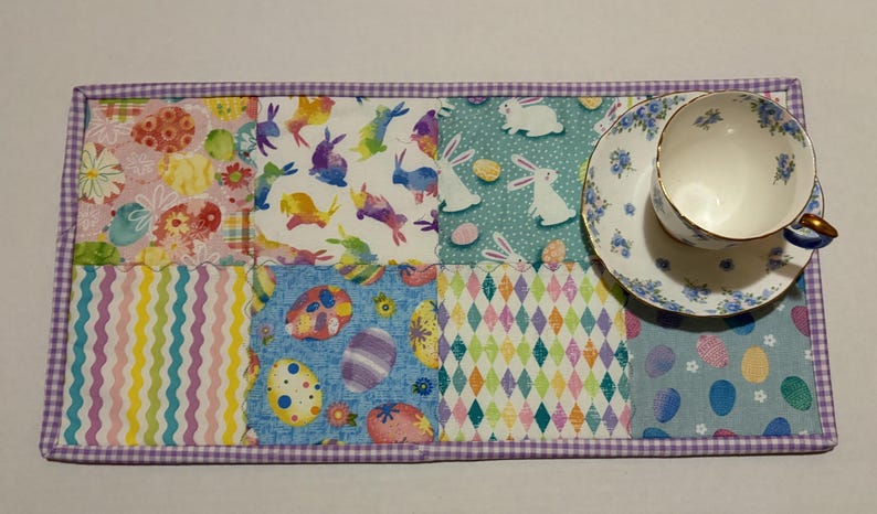 Może przedstawiać: Kolorowa podkładka na st&oacute;ł w tematyce wielkanocnej z filiżanką i spodkiem. Podkładka ma kwadraty patchworkowe z pisankami, kr&oacute;liczkami i pastelowymi wzorami. Filiżanka i spodek mają niebieski kwiatowy wz&oacute;r.