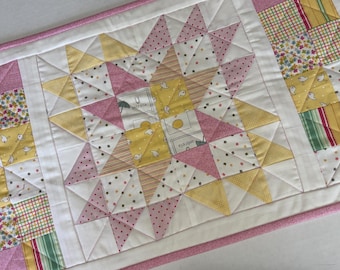 Peut inclure: Un chemin de table matelassé rectangulaire présentant un motif d'étoile central composé de triangles de tissu roses, jaunes et blancs. Le design inclut de petits motifs d'oiseaux et de parapluies, avec des bordures à carreaux et florales.