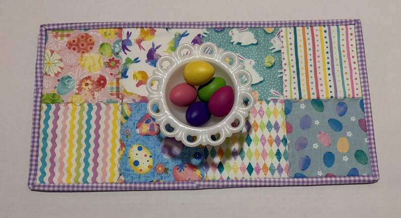 Może przedstawiać: Prostokątna podkładka pod talerz w tematyce wielkanocnej z patchworkowym wzorem z pastelowymi wzorami i białą, karbowaną miseczką wypełnioną kolorowymi jajkami. Podkładka ma fioletową obw&oacute;dkę w kratkę vichy.