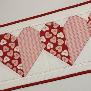 May include: A Valentine's Day themed table runner featuring alternating heart patterns. Some hearts are red with white stripes, others are red with white hearts and text. The runner has a white background with red trim.