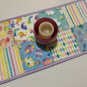 Może przedstawiać: Kolorowy bieżnik wielkanocny z patchworkowym wzorem. Bieżnik przedstawia r&oacute;żne wzory, w tym pisanki, kr&oacute;liki i kształty geometryczne. Na wierzchu znajduje się bordowy świecznik ze świecą. Krawędzie obszyte są fioletową kratką.