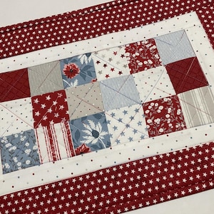 May include: A rectangular quilted placemat with a patchwork design. The center features squares of red, white, and blue fabrics with various patterns, including stars, stripes, and floral prints. The border is red with white stars.