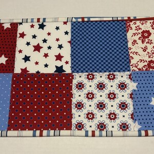 May include: A rectangular patchwork fabric item with a patriotic theme. It features eight squares with various patterns: red with white stars, white with red and blue stars, blue gingham, red floral, blue with white dots, red with white dots, white with red floral, and blue with white stars.